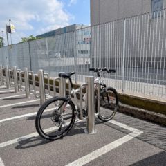 Nell'immagine, vediamo un'area di parcheggio per biciclette attrezzata con dissuasori fissi Pilomat 140-900 LH in acciaio inox satinato. Una bicicletta è assicurata a uno dei dissuasori, evidenziando la praticità e la sicurezza di questi dispositivi.