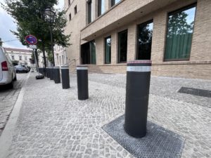 Row of Pilomat automatic anti-terrorism bollards with brushless electromechanical motor installed on an urban sidewalk in front of a brick building.